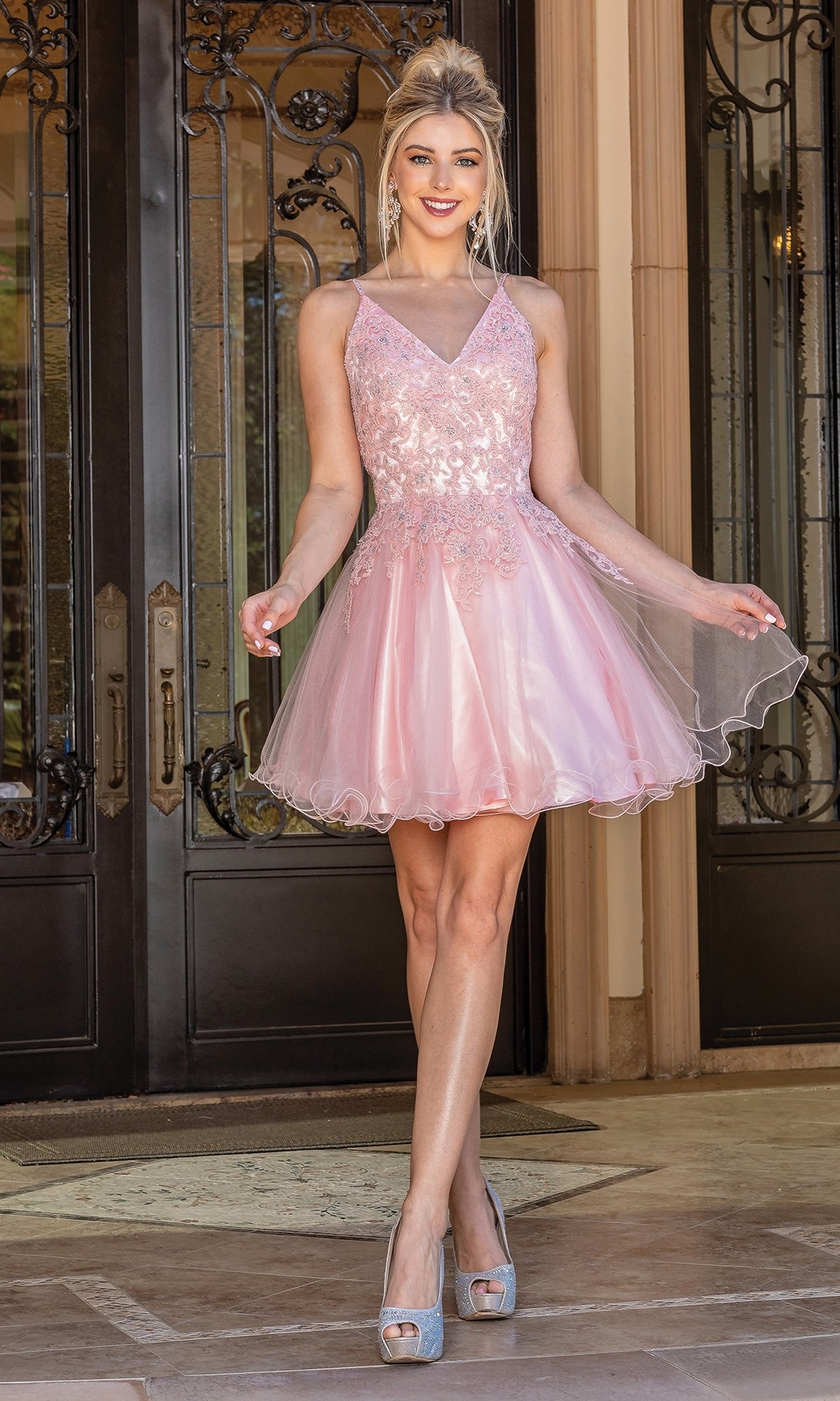 A woman stands before ornate doors wearing the Lace-Up Short Babydoll Homecoming Dress 3303, a sleeveless light pink dress with a tulle skirt and sequin details, paired with silver high heels.